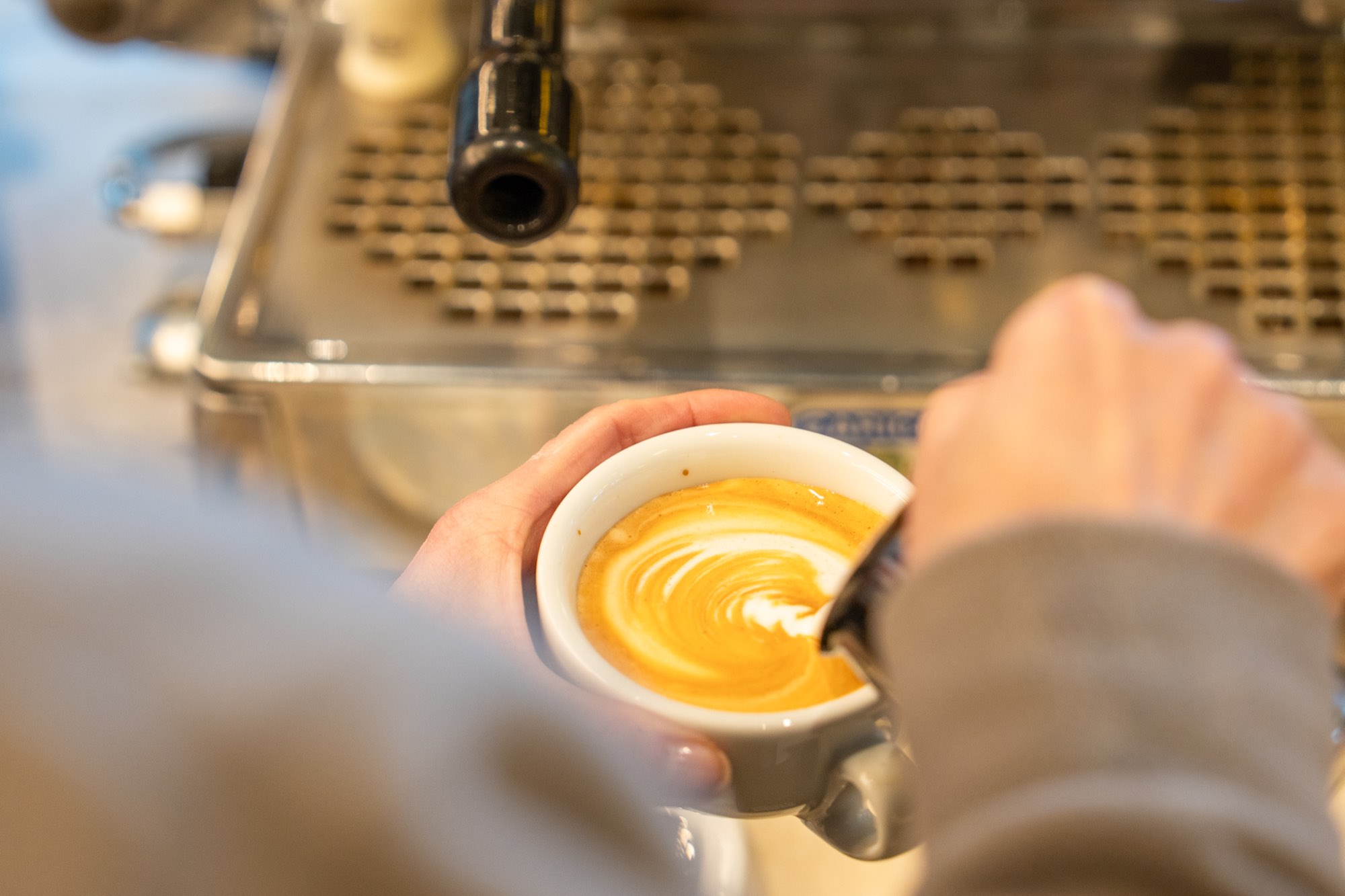 Barista gießt Milchschaum aus dem Milchkännchen in einen Espresso und formt Latte Art vor einer Siebträgermaschine