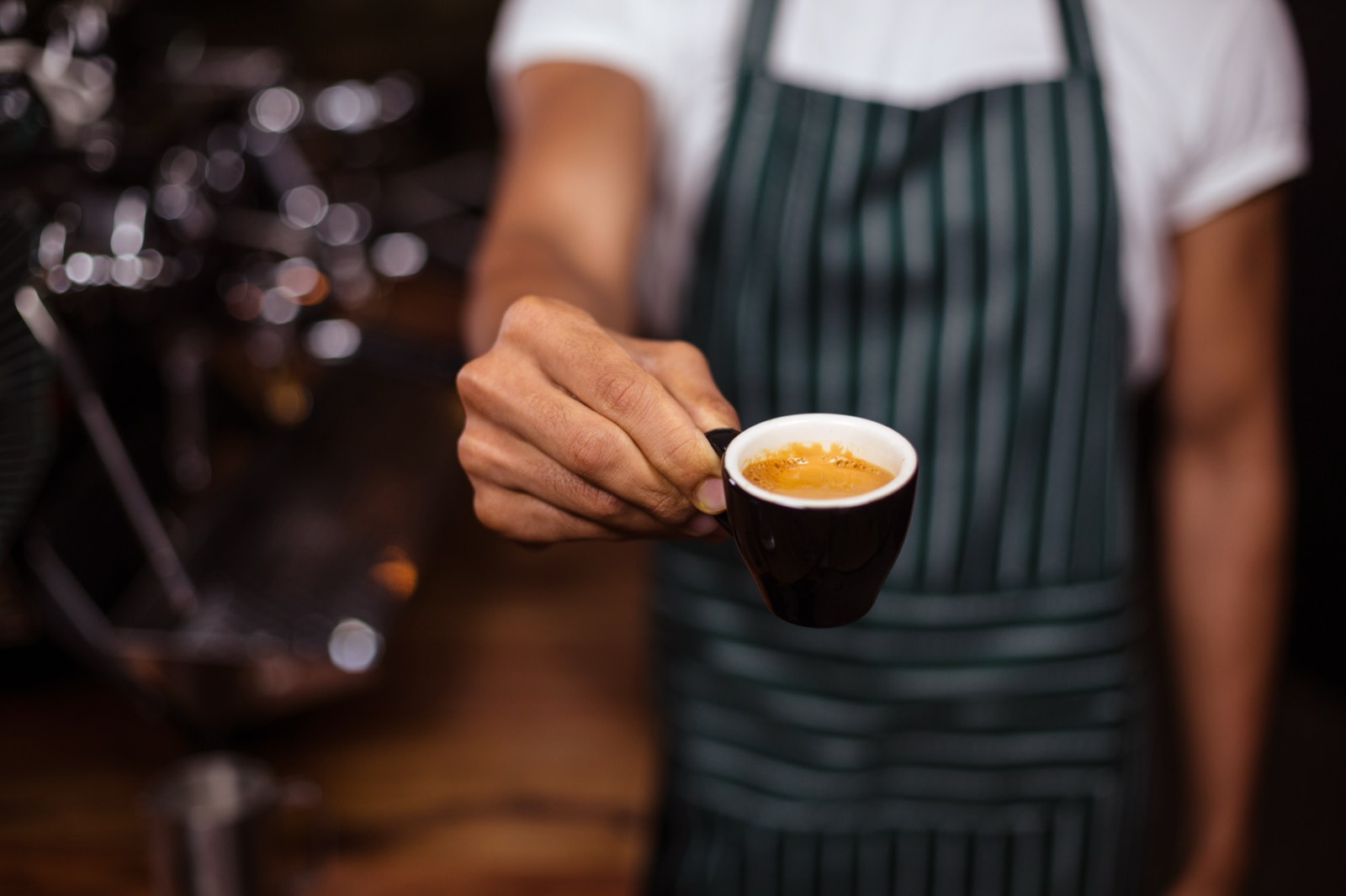 Barista in gestreifter Schürze reicht eine Tasse Espresso mit goldbrauner Crema