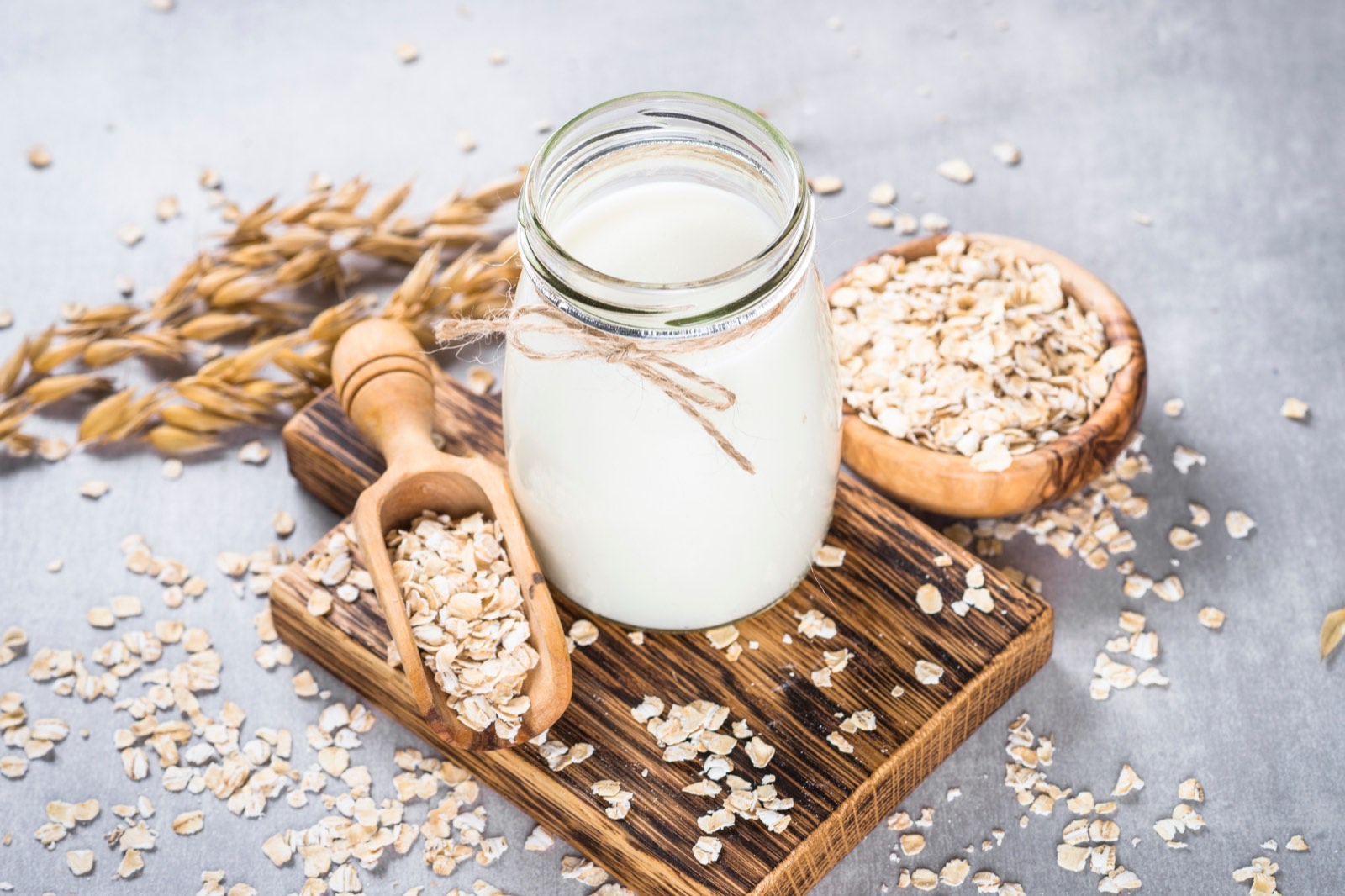 Oat milk in a glass with a string, next to it oatmeal, oat ears, and a wooden ladle