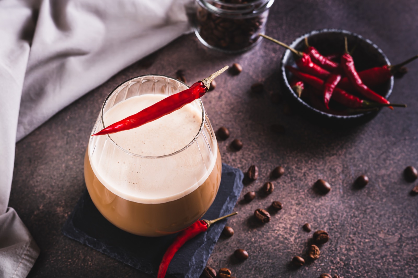 Coffee with foamed milk in a glass, garnished with a red chili pepper, with coffee beans and a bowl of chili peppers next to it