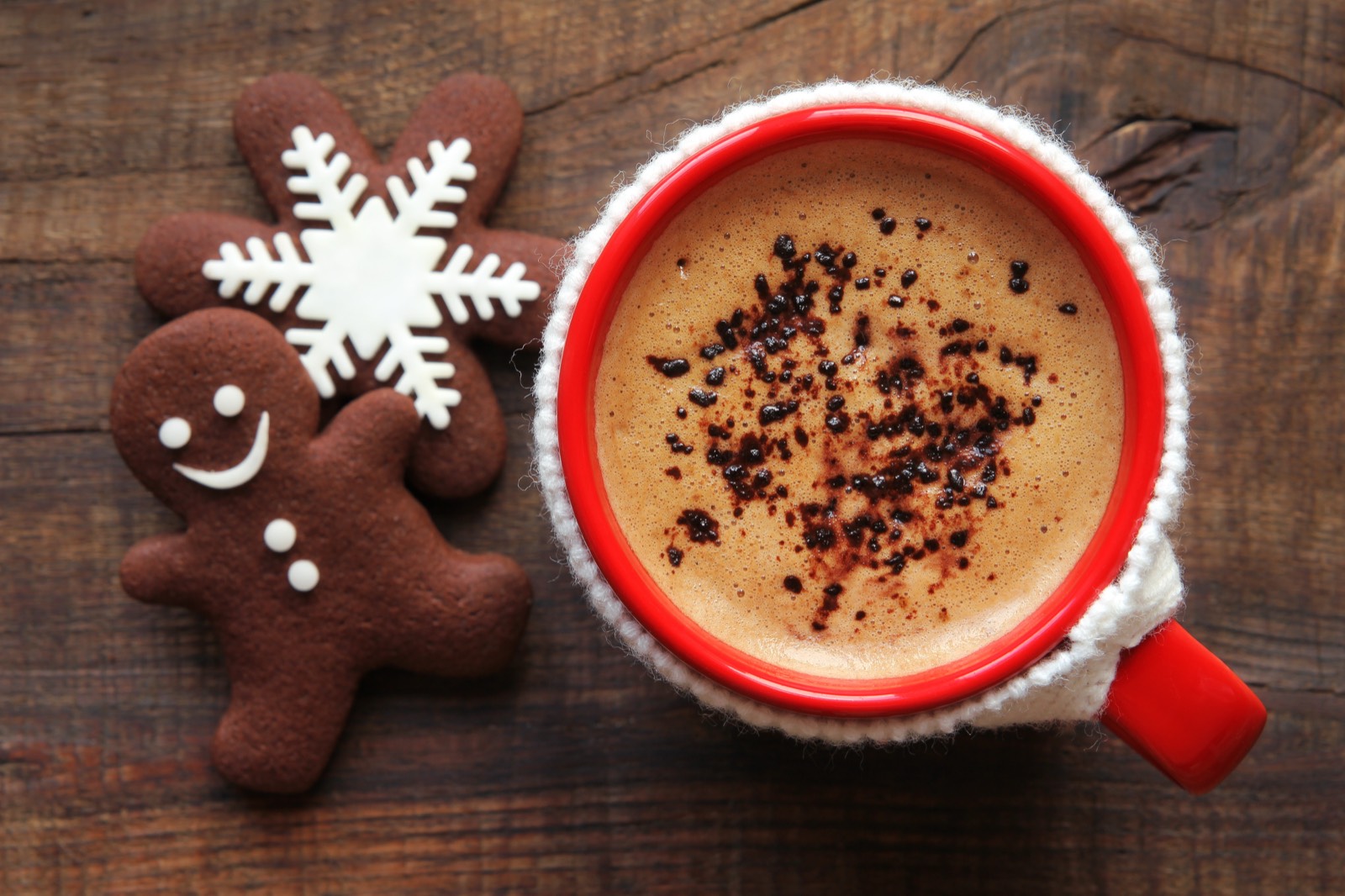 Coffee with foamed milk and cocoa powder in a red mug with a knitted sleeve, next to it a gingerbread man and a snowflake cookie