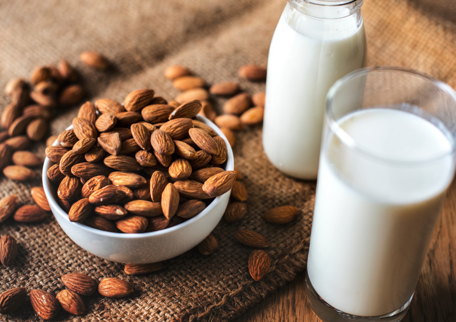 Almond milk in a glass and a bottle, next to a bowl of almonds on a jute mat